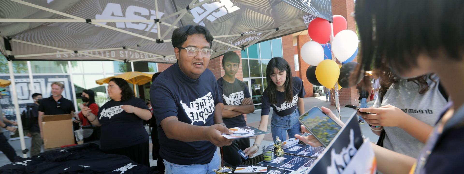一名学生穿着印有“我们人民”字样的t恤，站在联合学生公司下面.，林冠递给学生一本小册子.